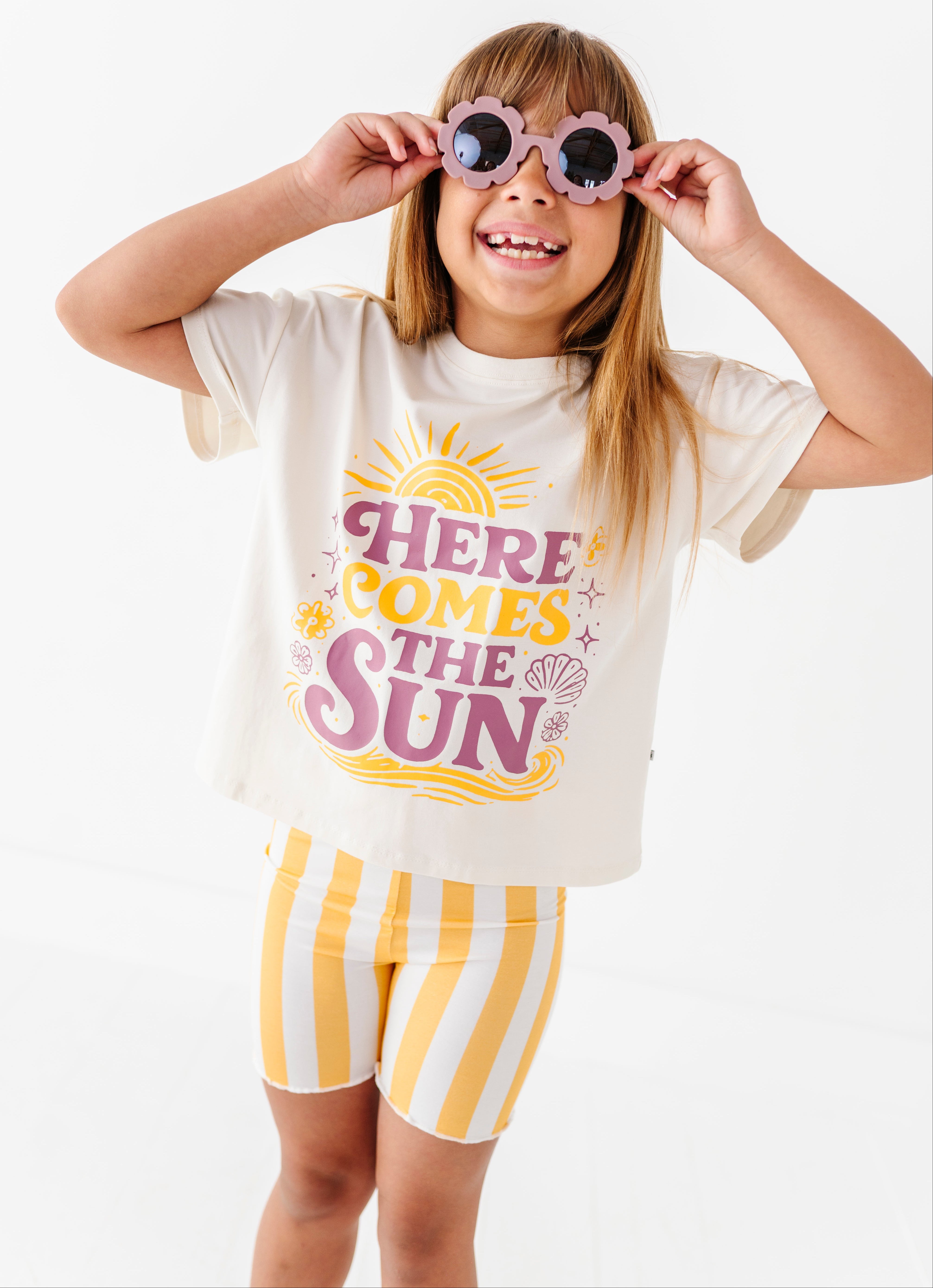 Child wearing a 'Here Comes the Sun' t-shirt and yellow striped shorts on a white background